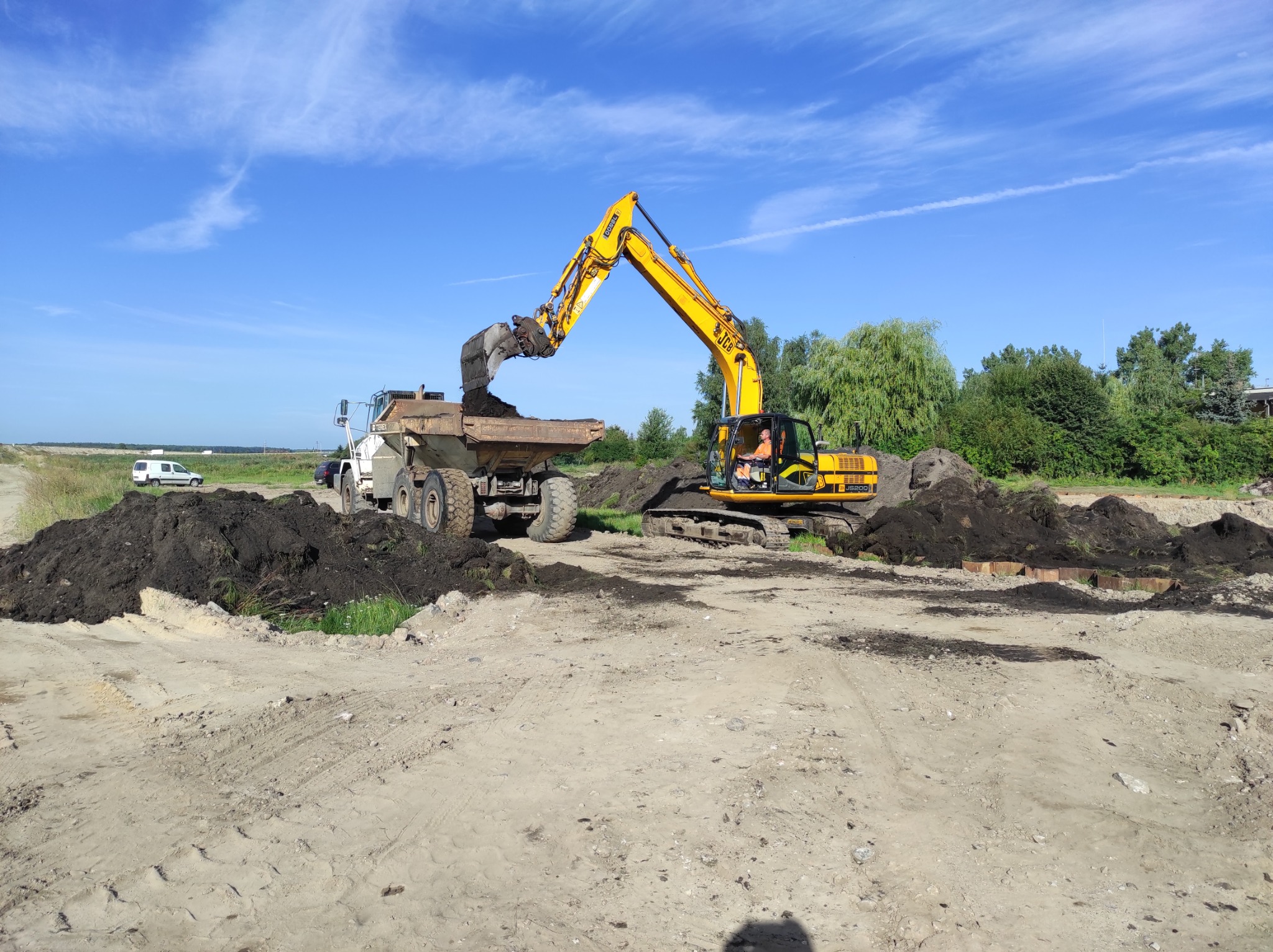 Żółta koparka JCB JS200 nabiera ziemię i wysypuje ją do wywrotki na tle błękitnego nieba z delikatnymi chmurami.