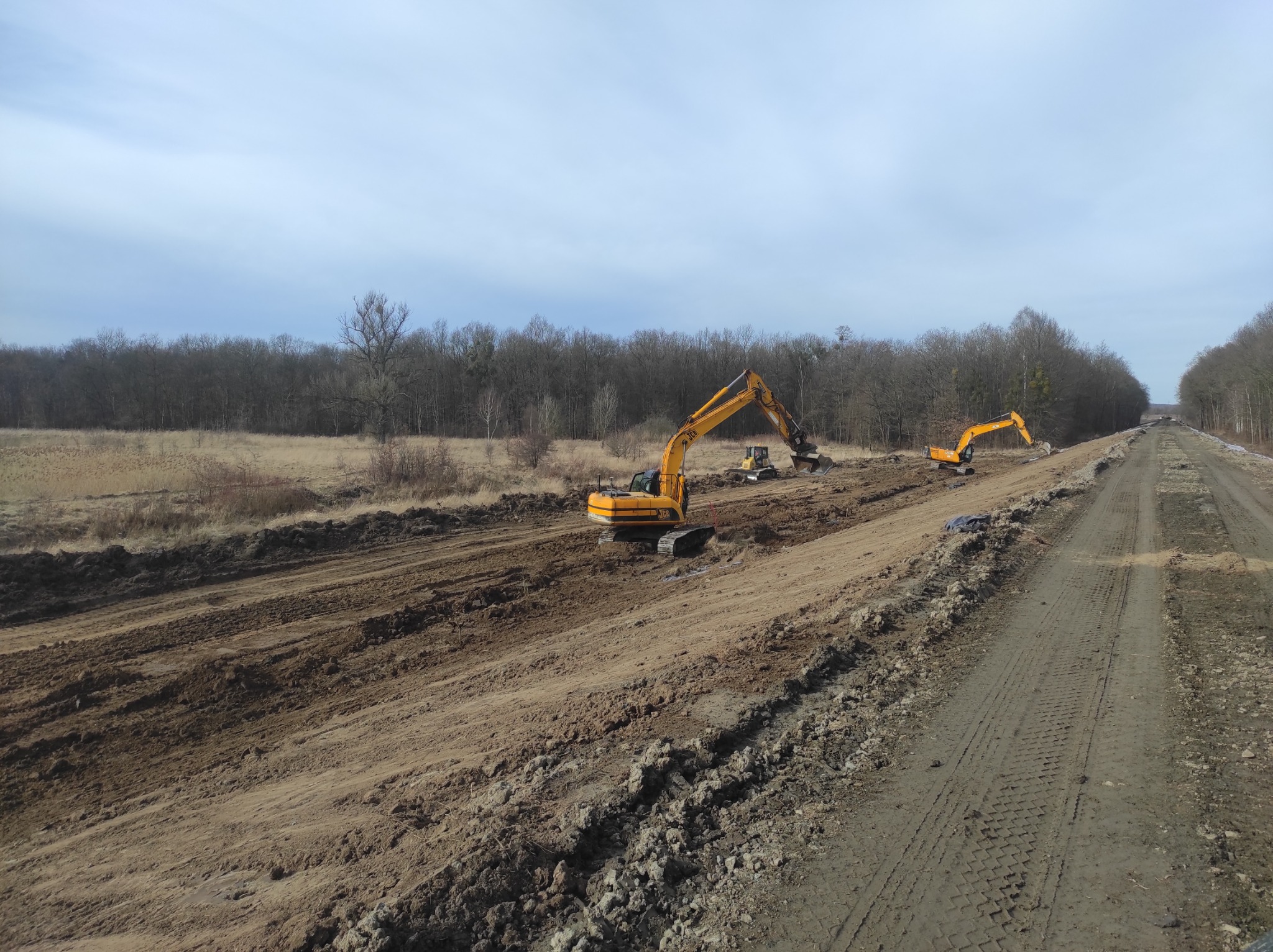 Żółte koparki pracują na placu budowy, przygotowując teren pod drogę; w tle widoczny las bez liści.