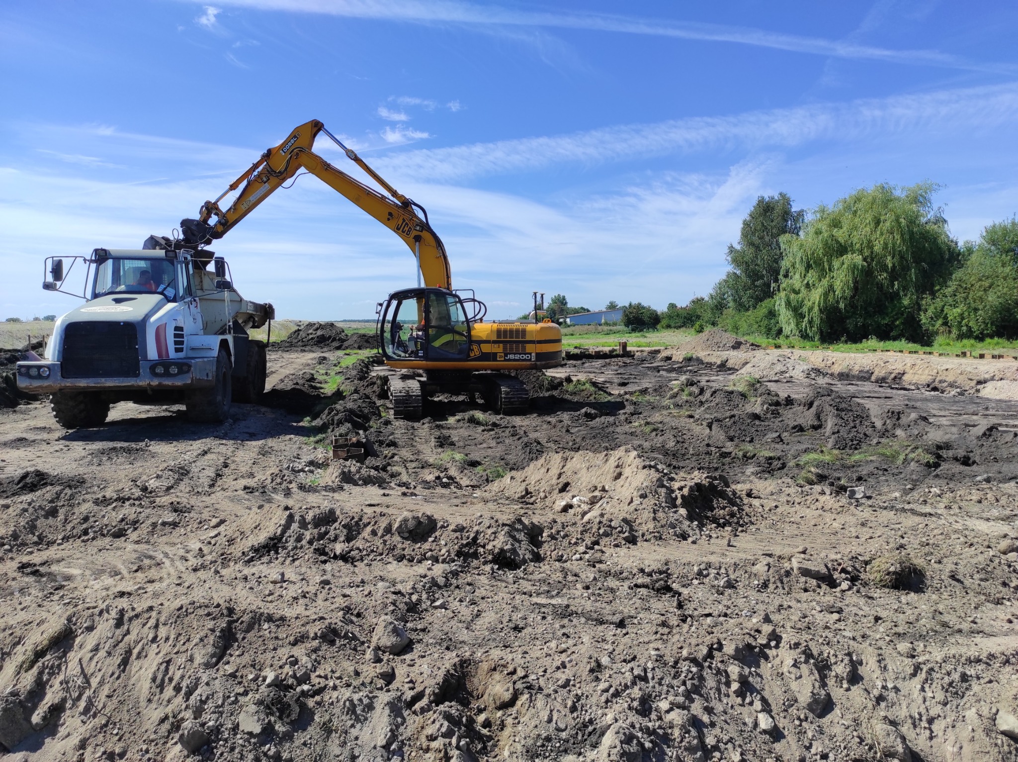 Żółty koparka JCB JS200 i biała wywrotka Combel podczas prac ziemnych na nierównym, przygotowanym terenie pod budowę, błękitne niebo z chmurami w tle.