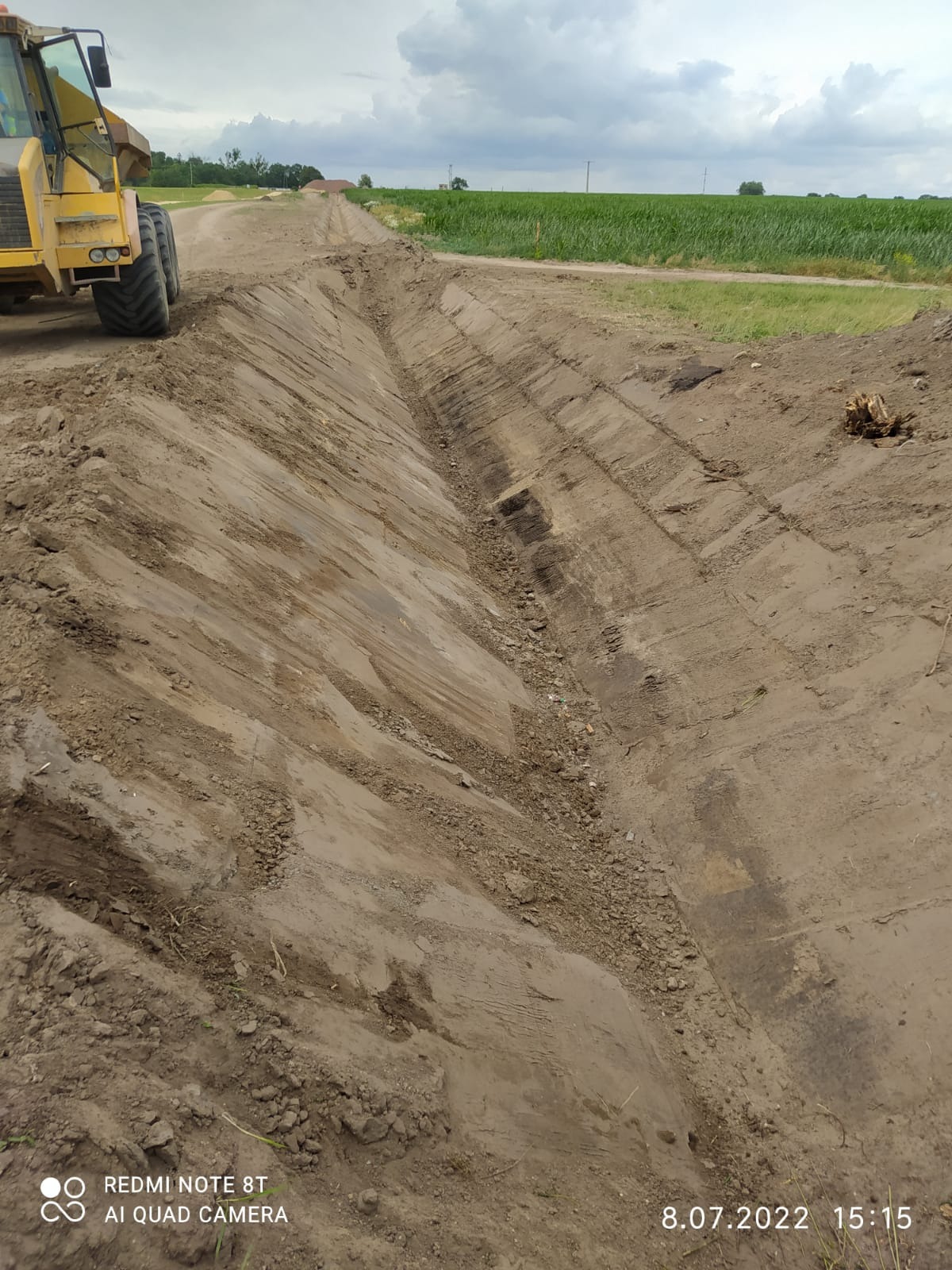 Żółta wywrotka częściowo widoczna przy wykopanym rowie melioracyjnym na polu, w tle zielone uprawy i pochmurne niebo.
