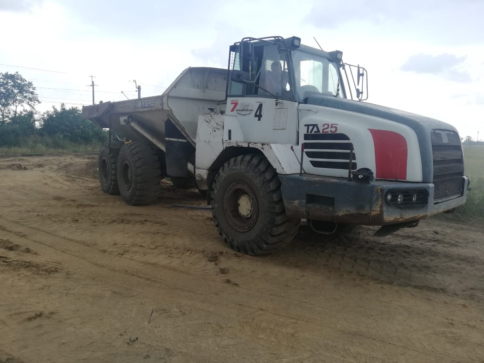 Wyładowany wozidło technologiczne TA25 na piaszczystym terenie budowy, widoczne ślady opon i fragmenty zieleni w tle.