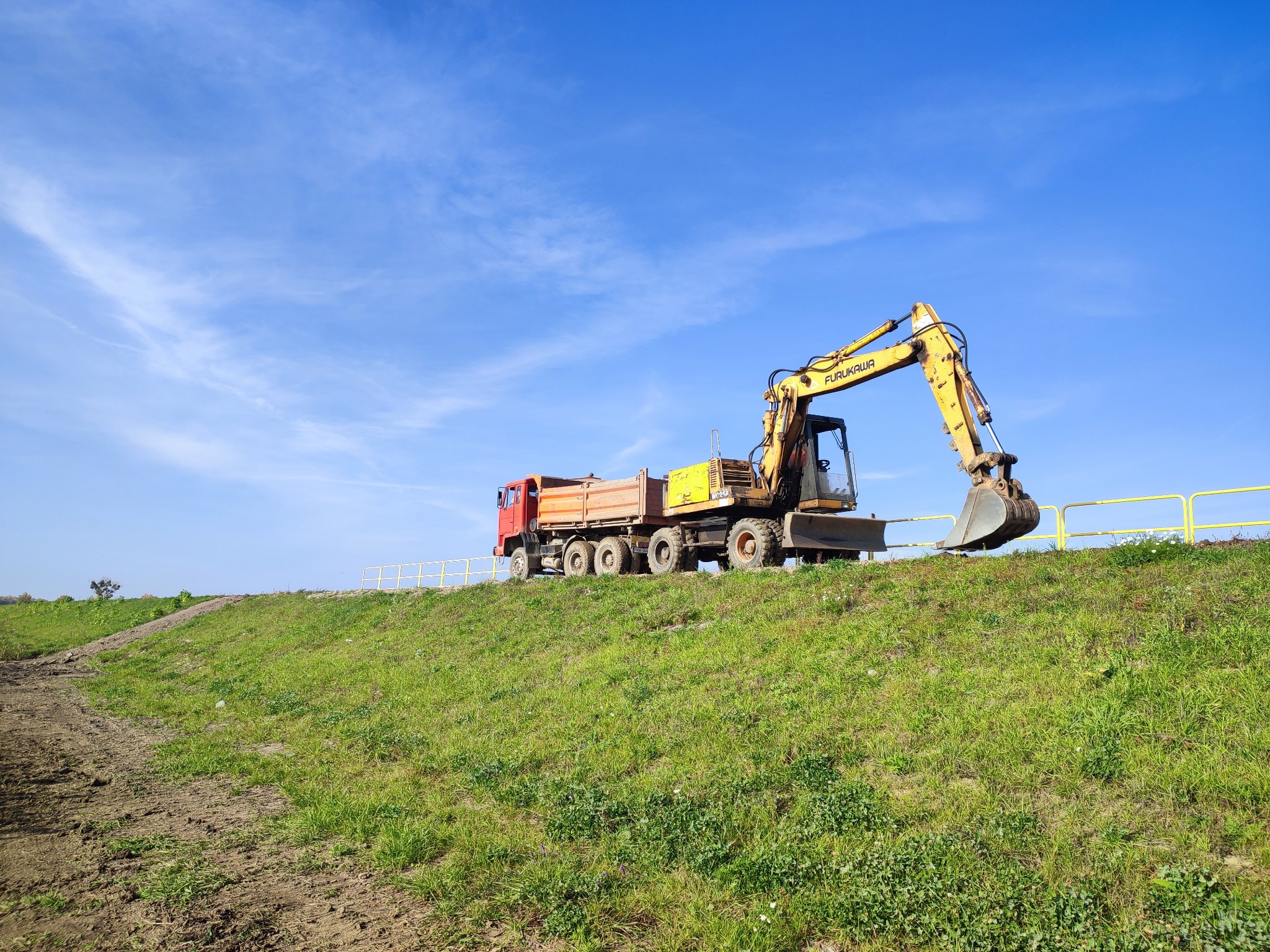 Żółta koparka Furukawa i czerwona wywrotka na szczycie zielonego wzgórza pod błękitnym niebem z lekkimi chmurami.