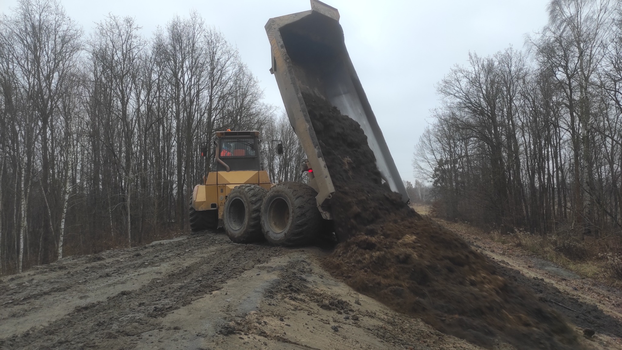 Żółta wywrotka terenowa wysypuje ziemię na nierówny, błotnisty grunt na tle lasu z nagimi drzewami w pochmurny dzień.