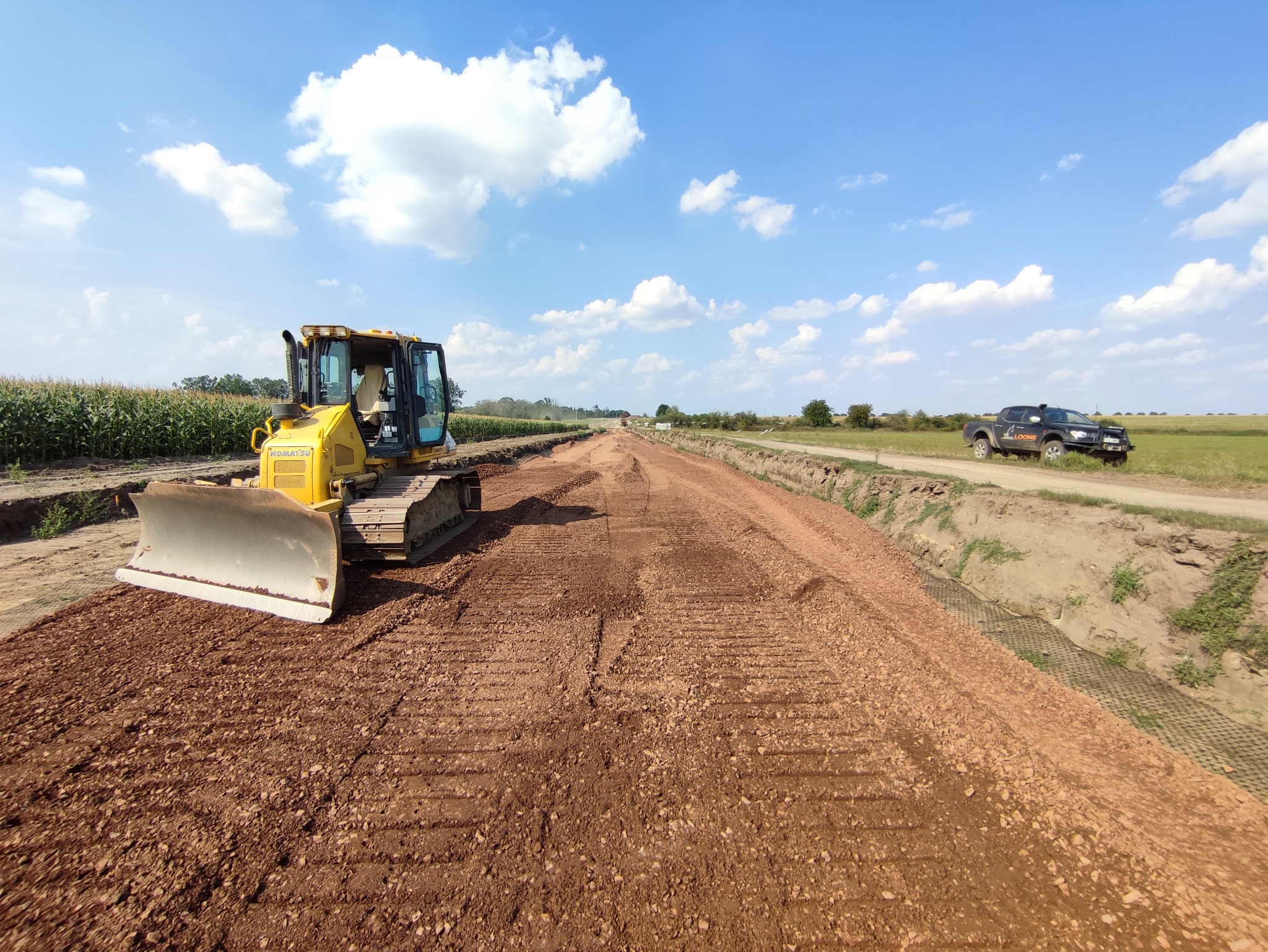 Żółty spychacz Komatsu wyrównuje czerwoną, piaszczystą nawierzchnię przyszłej drogi, z widocznymi śladami gąsienic i lemiesza, w tle pole kukurydzy i samochód terenowy.
