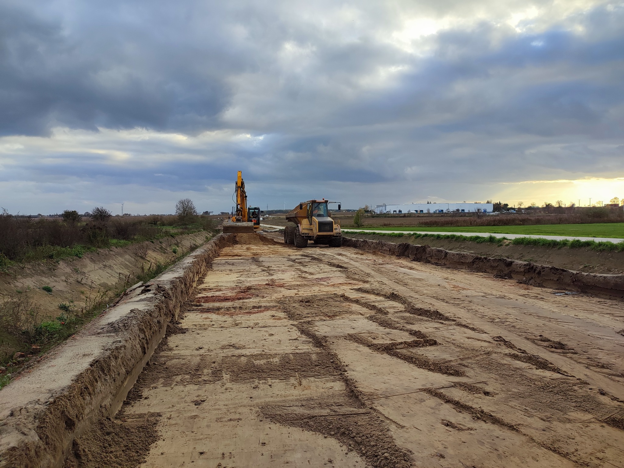 Przygotowanie podłoża pod drogę z widocznym wykopem, utwardzonym gruntem, koparką i wywrotką na tle pochmurnego nieba.