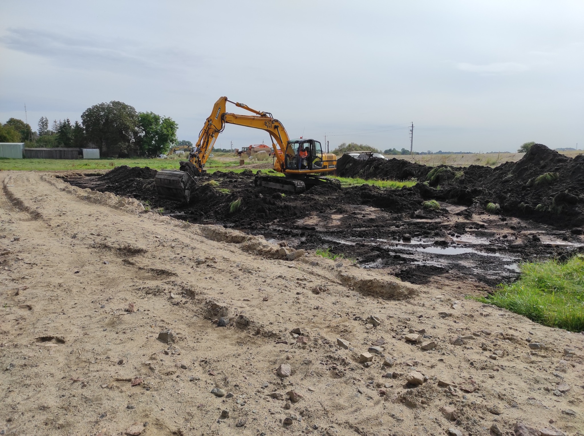 Żółta koparka JCB na nierównym, piaszczystym terenie z wykopanymi rowami i stertami ziemi, w tle widoczne budynki gospodarcze i drzewa pod pochmurnym niebem.