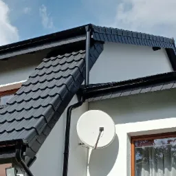 Nowoczesny dom z czarnym dachem z blachodachówki, czarnymi rynnami i białą elewacją, antena satelitarna na ścianie.