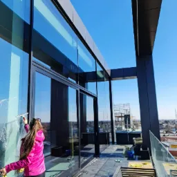 Kobieta w r&oacute;żowej kurtce czyści szklaną fasadę budynku za pomocą ściągaczki i ż&oacute;łtej ścierki, widok na panoramę miasta w tle.