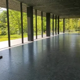 Wnętrze przestronnego pomieszczenia z betonową podłogą po umyciu, widoczne refleksy światła i resztki wody, duże okna z widokiem na zieleń drzew, w rogu leży szara ścierka.