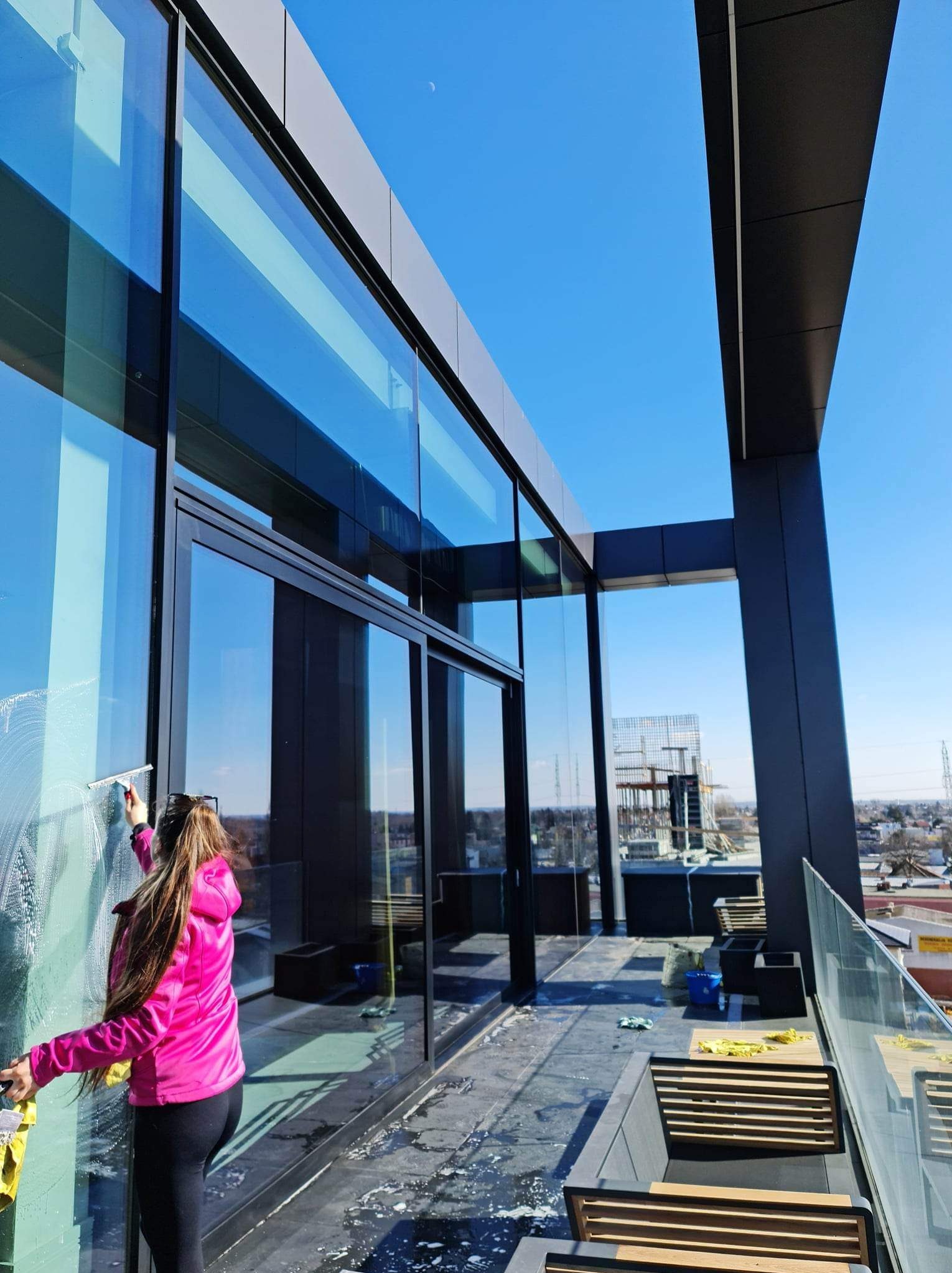 Kobieta w różowej kurtce czyści szklaną fasadę budynku za pomocą ściągaczki i żółtej ścierki, widok na panoramę miasta w tle.