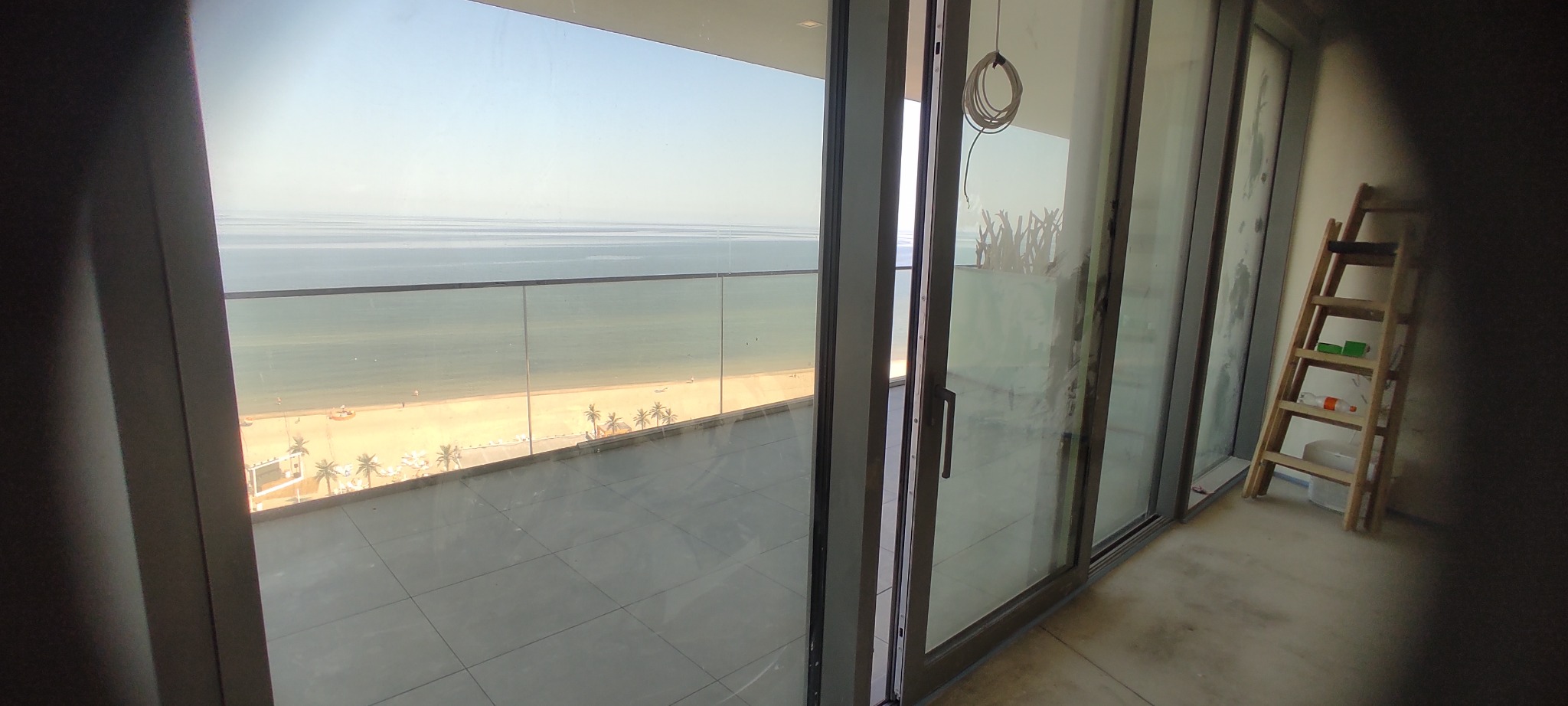 Widok z balkonu w Goleniowie na plażę i morze przez umyte, przesuwne okna. Na ścianie oparta drabina.