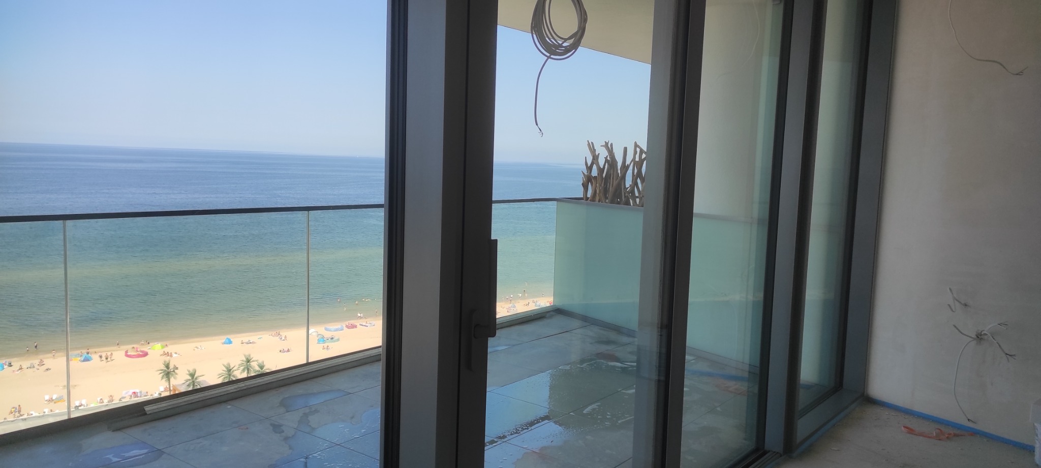 Widok z balkonu na plażę i morze przez przesuwne szklane drzwi; mokra podłoga balkonu po umyciu, widoczne niedokończone prace wykończeniowe na ścianie.