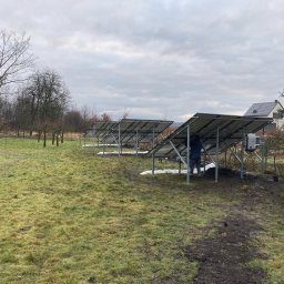 Apex Renewables Sp. z o.o. - Instalacja paneli słonecznych na ziemi w pochmurny dzień. Widoczny pracownik w niebieskiej kurtce i szarej czapce, montujący panele. Inwerter zamontowany na konstrukcji. W tle drzewa bez liści i dom.