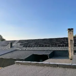 Instalacja fotowoltaiczna na dachu budynku przemysłowego z kominem. Panele ułożone rzędami, widok z góry na tle błękitnego nieba. Realizacja z wykorzystaniem OZE.