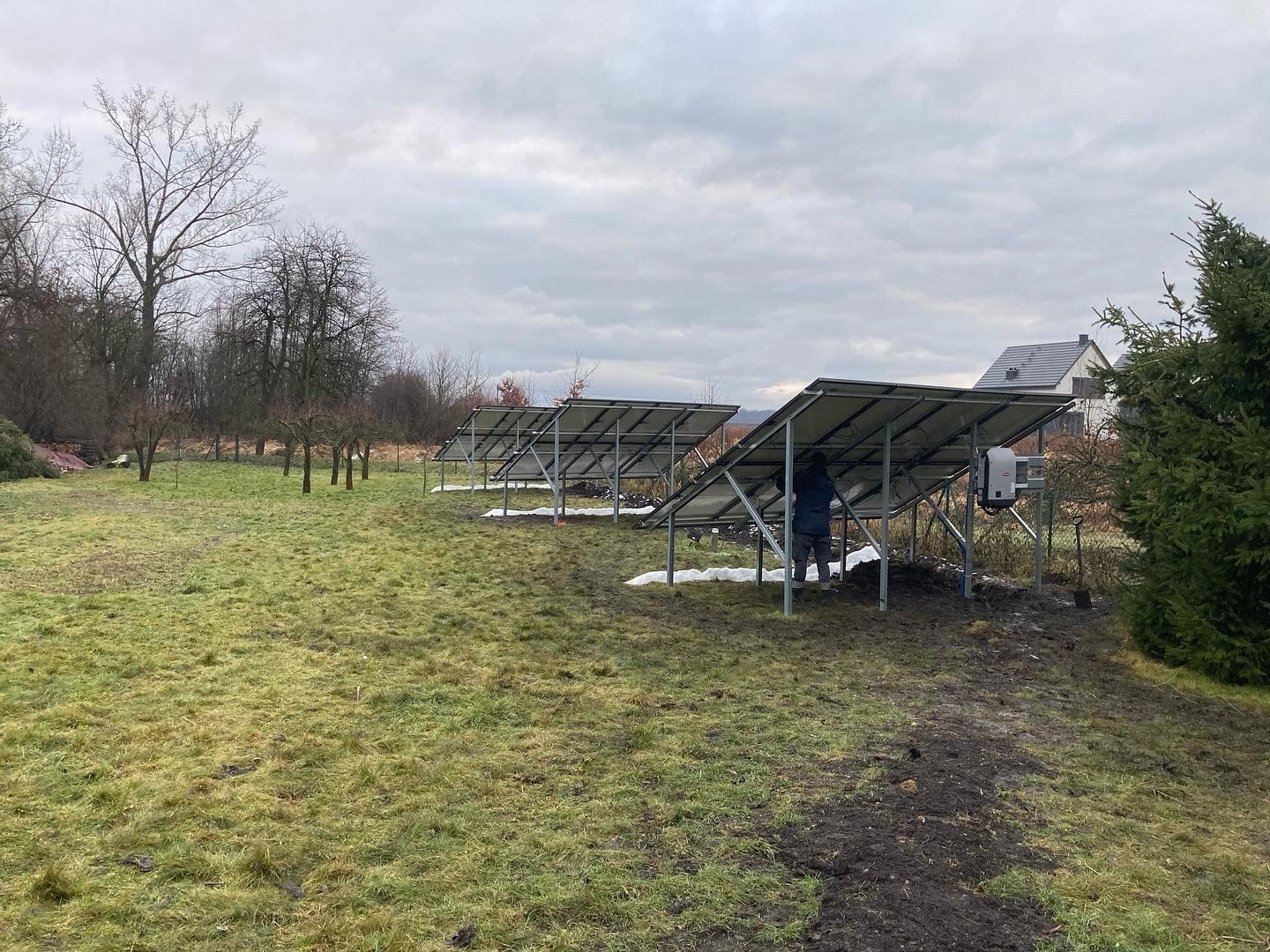 Instalacja paneli słonecznych na ziemi w pochmurny dzień. Widoczny pracownik w niebieskiej kurtce i szarej czapce, montujący panele. Inwerter zamontowany na konstrukcji. W tle drzewa bez liści i dom.