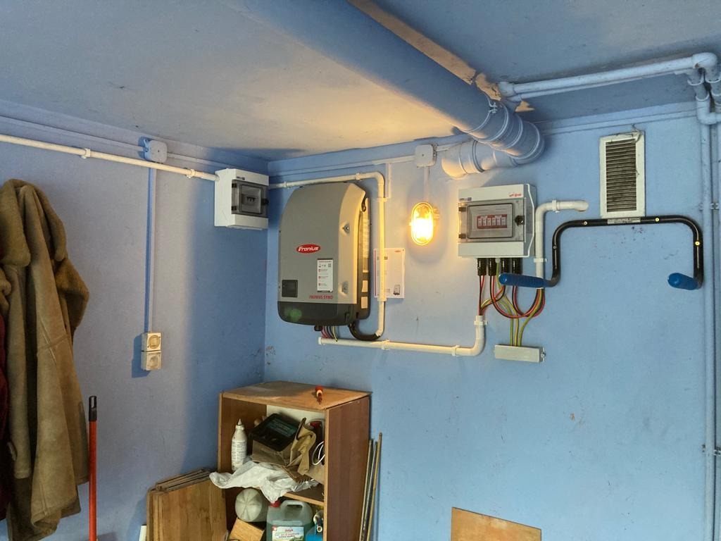 Szary falownik Fronius zamontowany na niebieskiej ścianie w garażu, obok skrzynki elektrycznej i rur instalacyjnych.