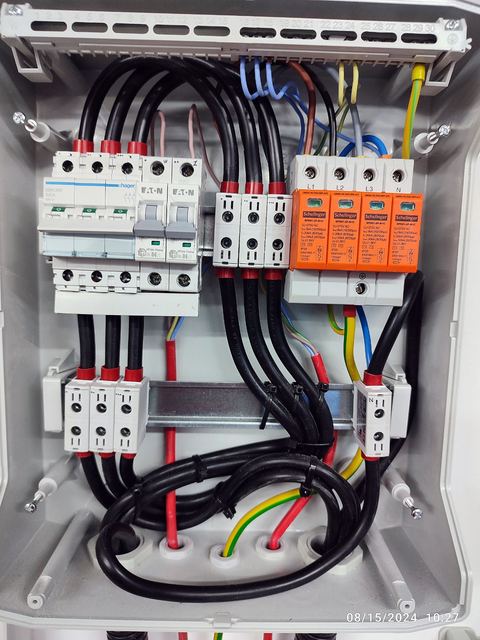 Szczegółowe ujęcie skrzynki elektrycznej z zainstalowanymi bezpiecznikami, przewodami i ogranicznikami przepięć. Widoczne elementy Hager, Eaton i Schellinger.