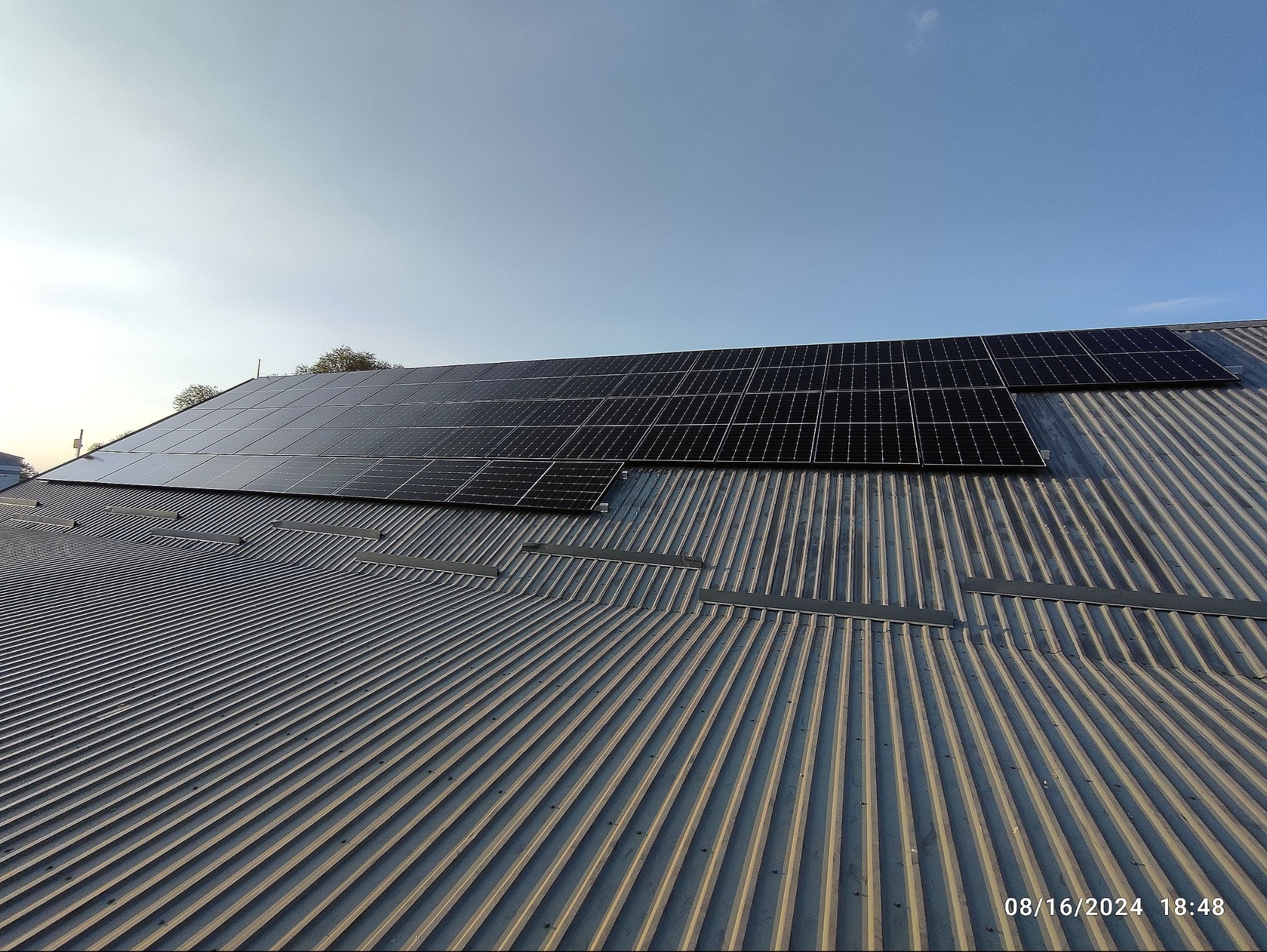 Panele słoneczne na metalowym, pofalowanym dachu budynku, widoczne z dołu pod kątem. Błękitne niebo w tle, delikatne oświetlenie, data i godzina w prawym dolnym rogu.