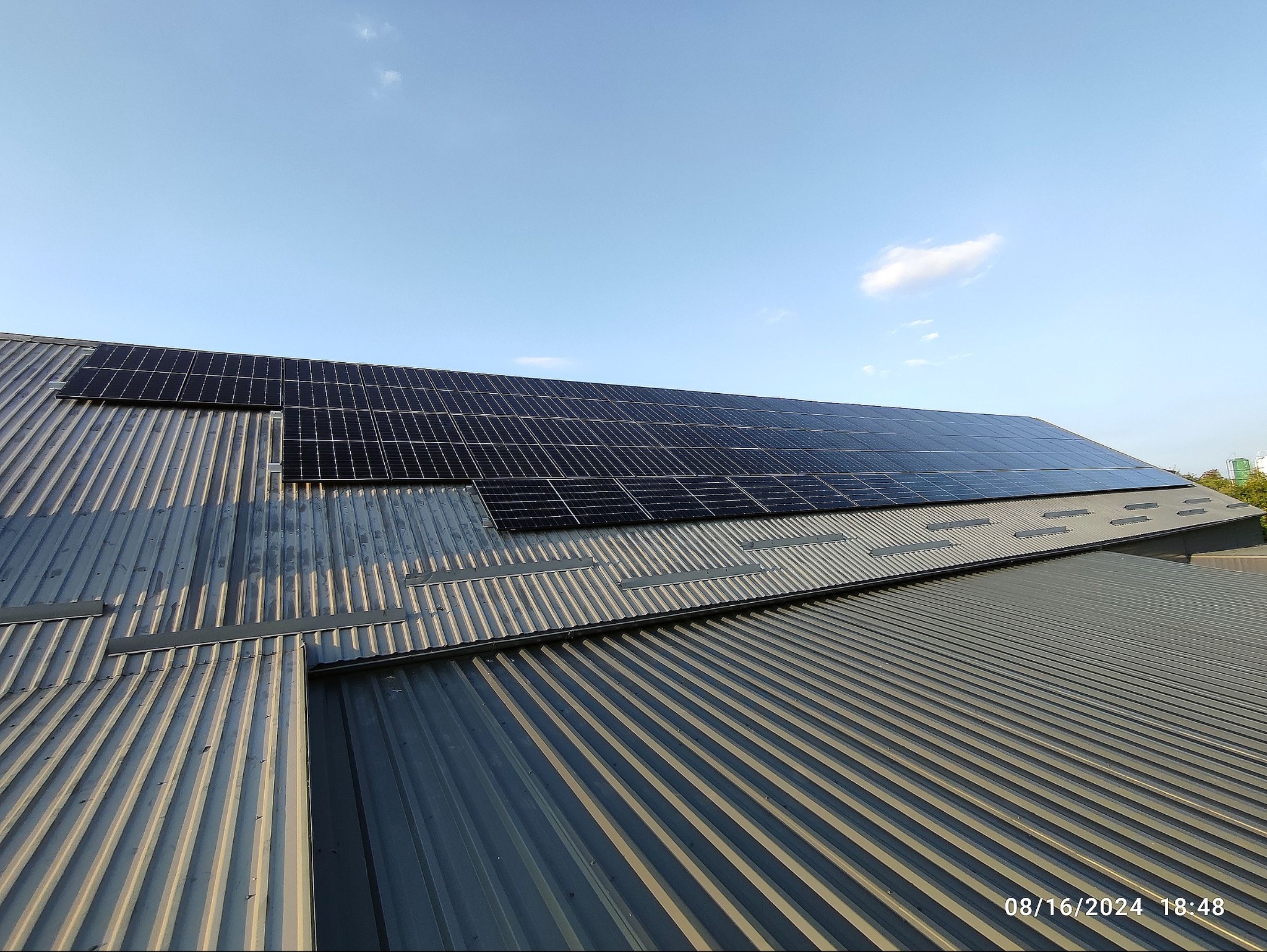 Instalacja paneli słonecznych na metalowym dachu budynku przemysłowego w słoneczny dzień. Widok z góry ukazuje panele i strukturę dachu.