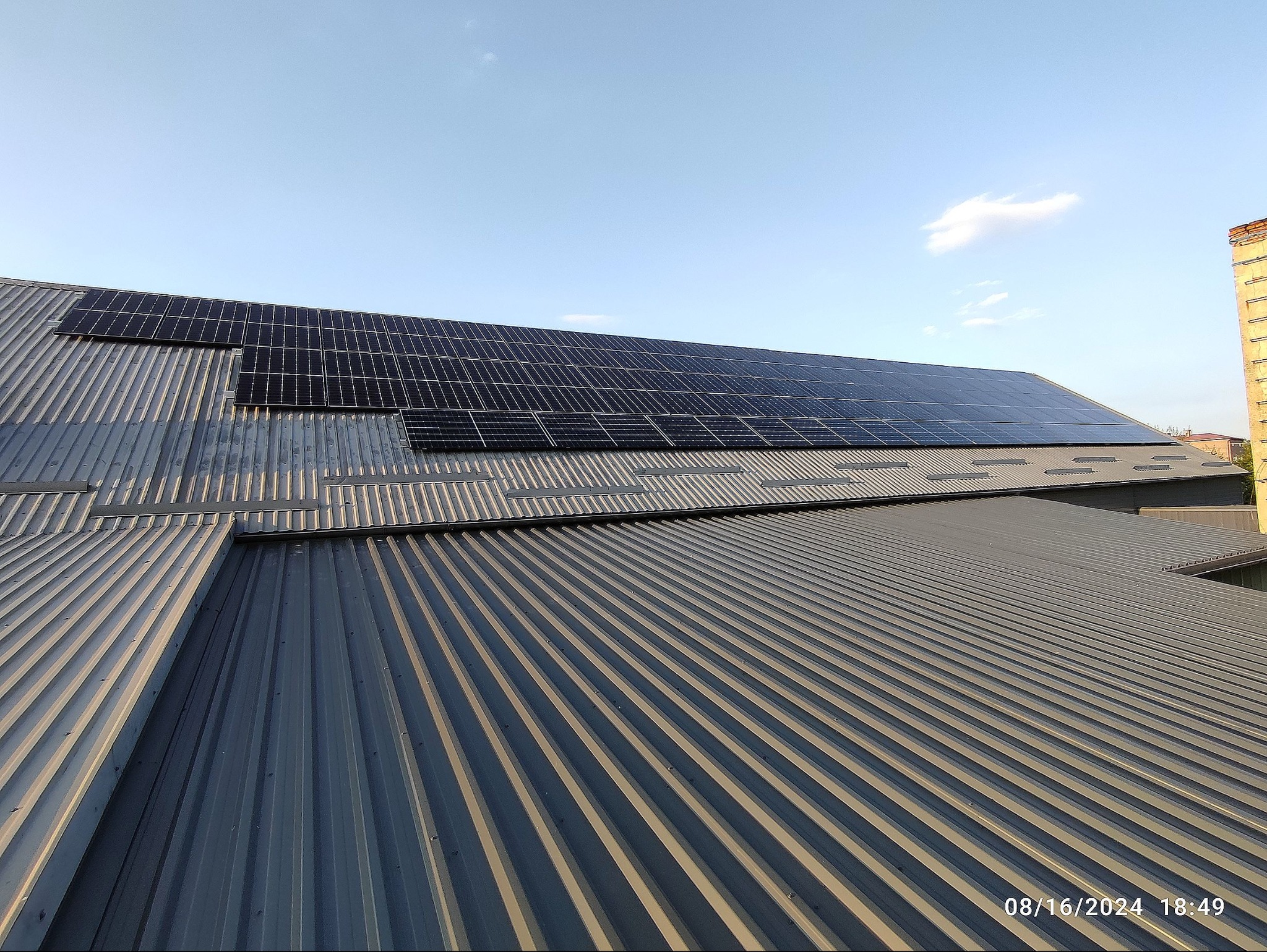 Panele fotowoltaiczne na falistej, blaszanej konstrukcji dachu przemysłowego budynku, widziane pod kątem, z fragmentem komina w tle i błękitnym niebem.