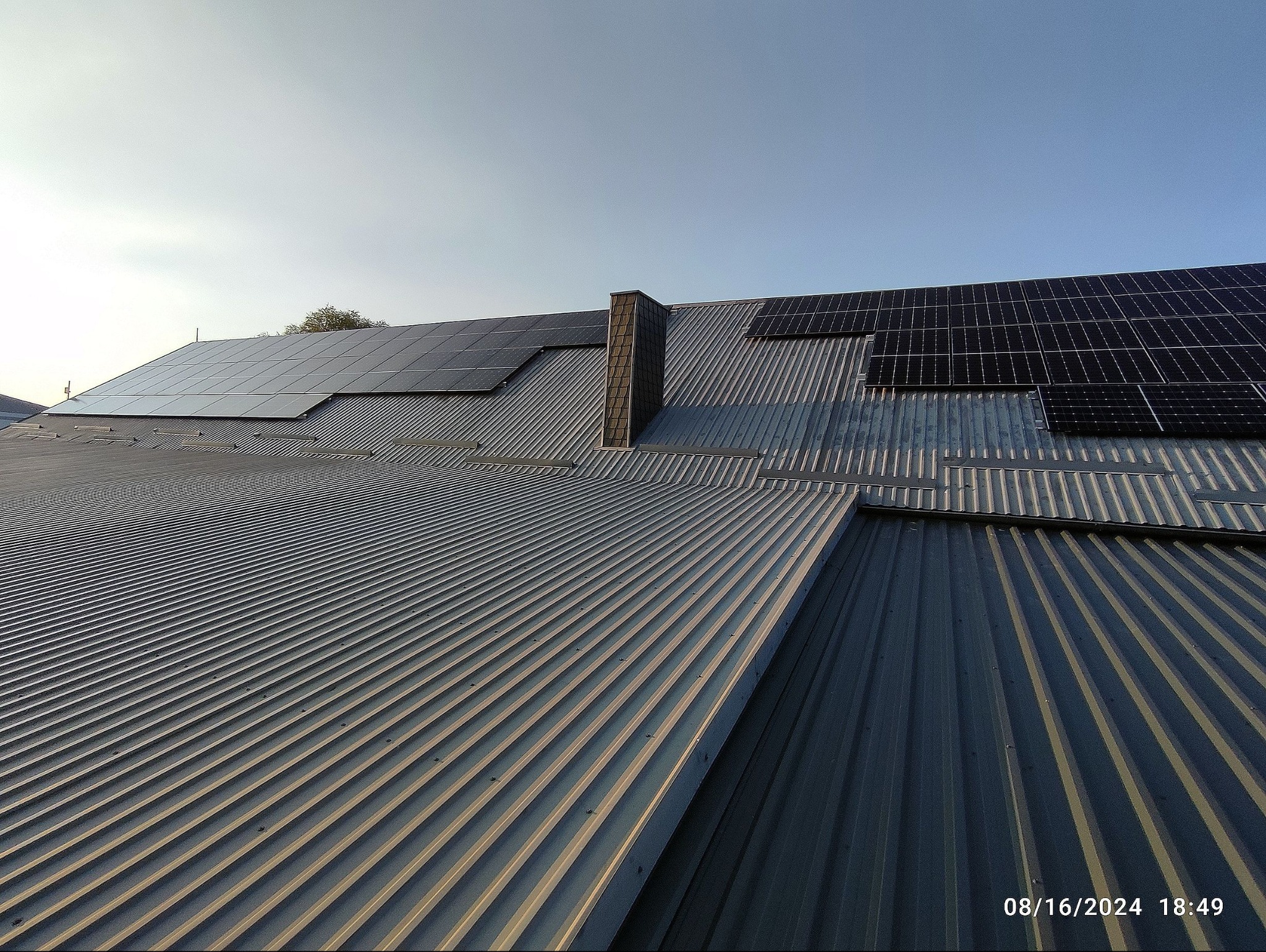 Instalacja paneli słonecznych na blaszanym dachu budynku we Wrocławiu. Widoczne panele i komin. Ujęcie z dołu, niebo w tle.