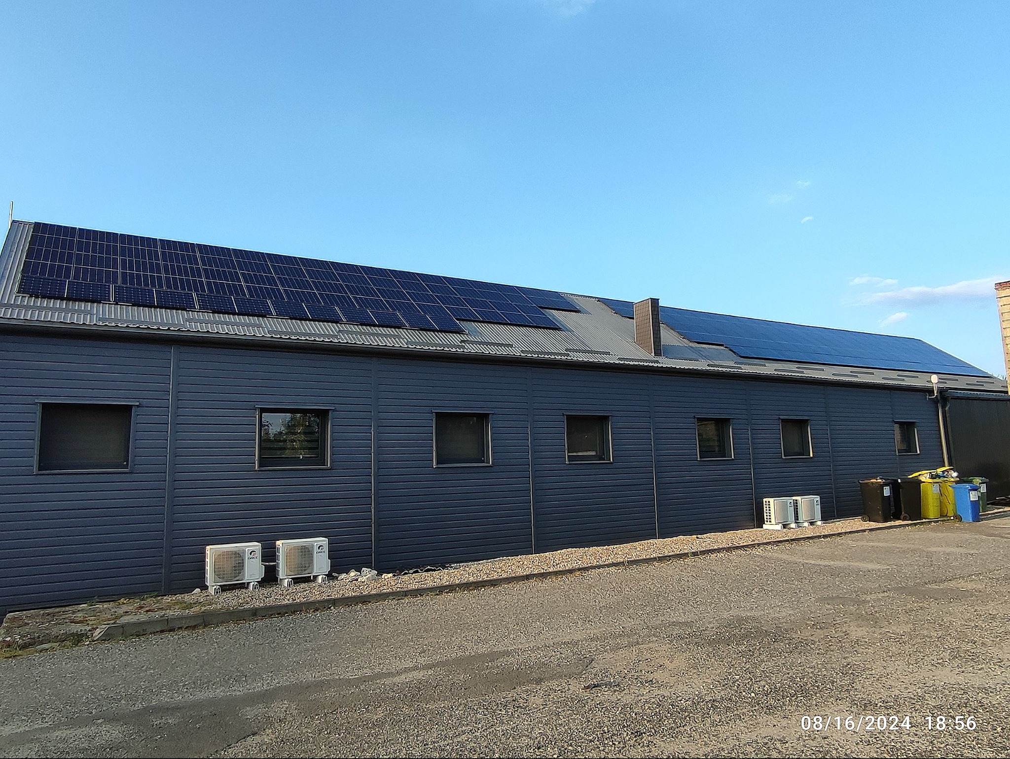Instalacja fotowoltaiczna na dachu budynku z szarej blachy falistej. Widoczne panele, komin i okna. Na dole trzy klimatyzatory i kosze na śmieci.