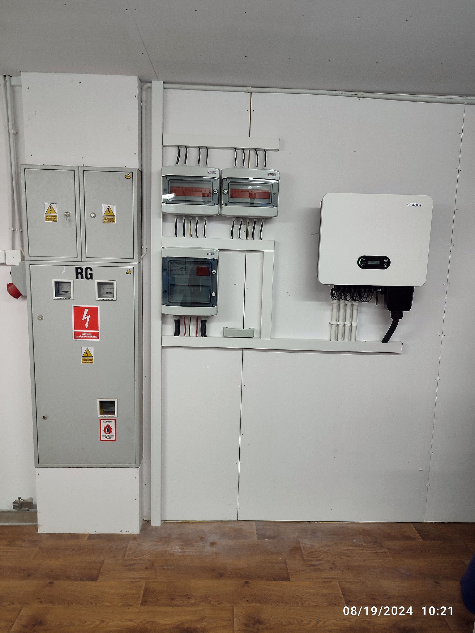 Szara skrzynka elektryczna RG, rozdzielnice z bezpiecznikami i biały inwerter SOFAR na białej ścianie z panelami. Instalacja elektryczna w pomieszczeniu.