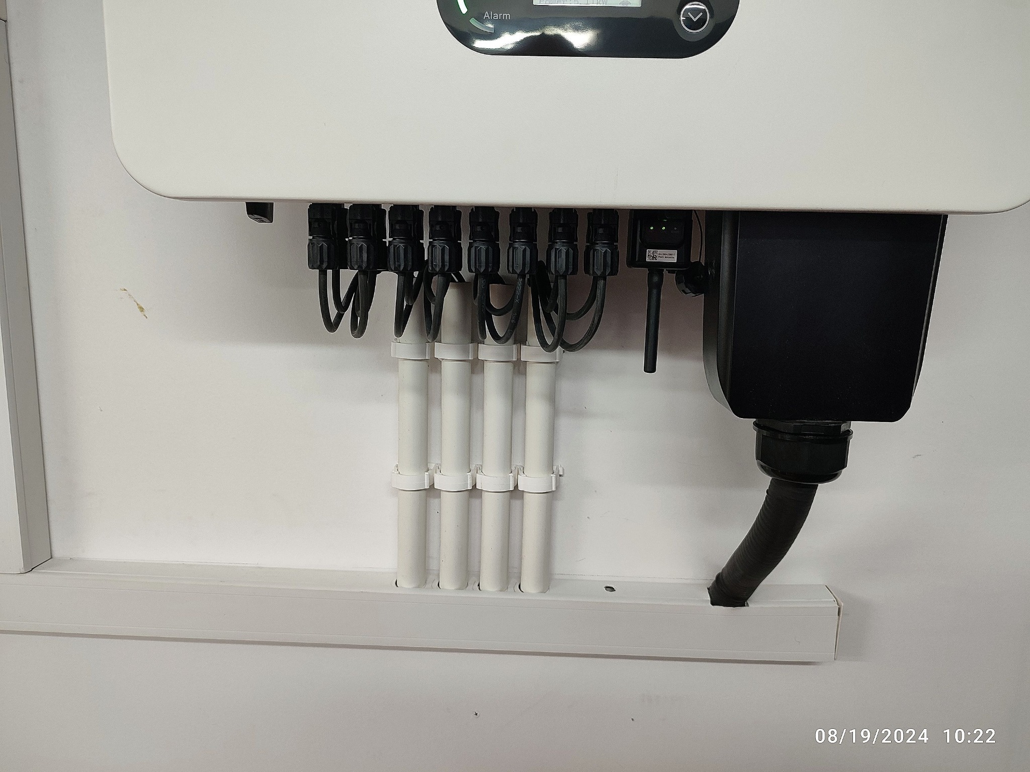 Szczegółowe ujęcie podłączenia przewodów do białego inwertera, z widocznymi czarnymi złączkami i białymi rurkami mocowanymi do ściany. Widoczne kable i obudowa.