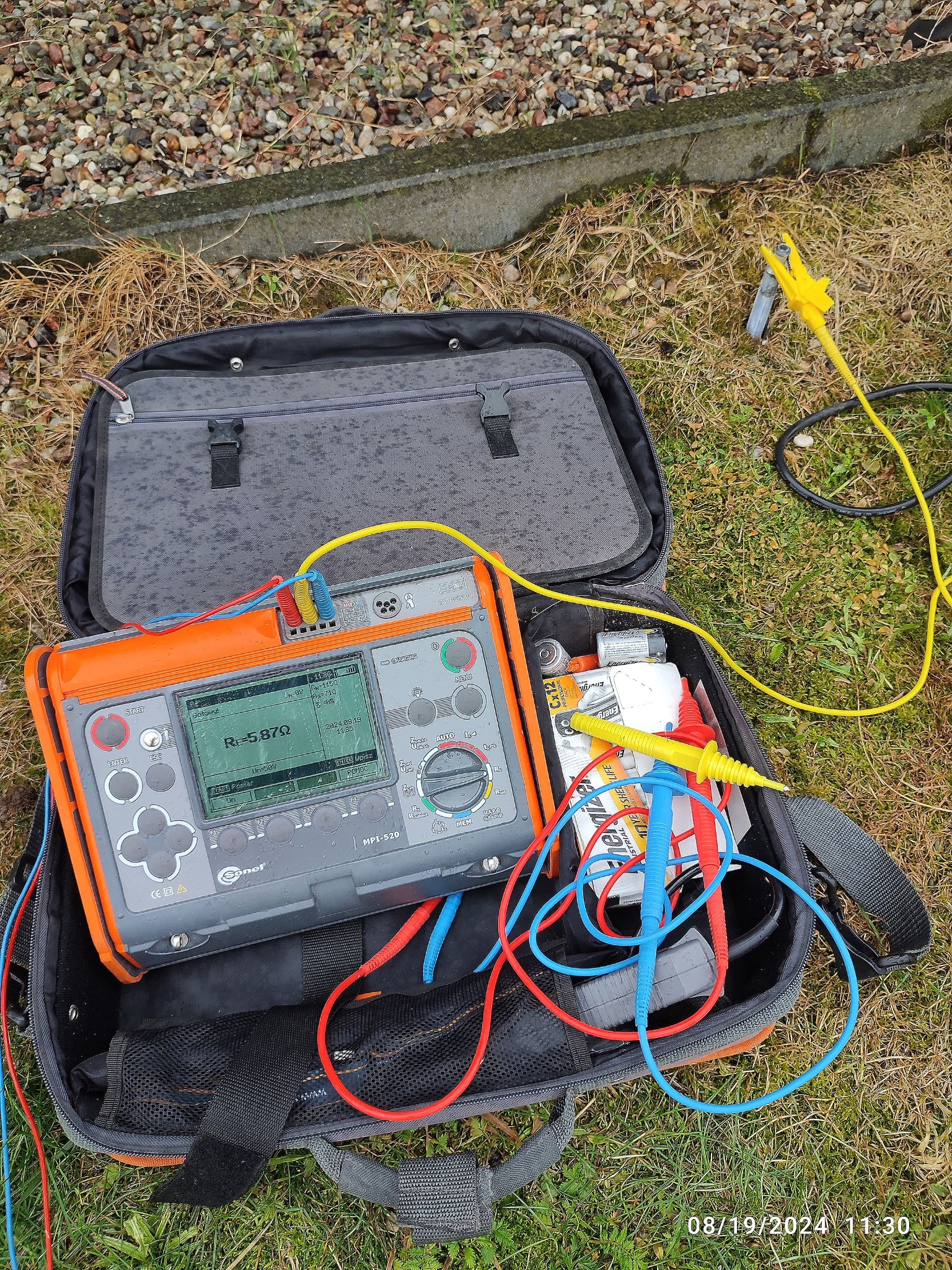 Miernik rezystancji uziemienia Sonel MPI-520 w otwartej torbie na trawie, z przewodami i krokodylkami. Wyświetlacz pokazuje R=5.87Ω. Widoczna data 2024.08.19.