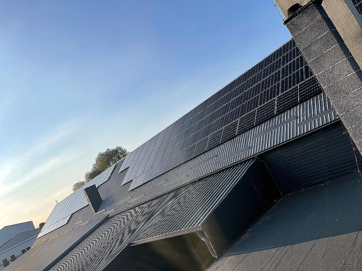 Instalacja fotowoltaiczna na dachu budynku z blachy trapezowej, widok pod kątem, błękitne niebo w tle. Panele w układzie rzędowym.