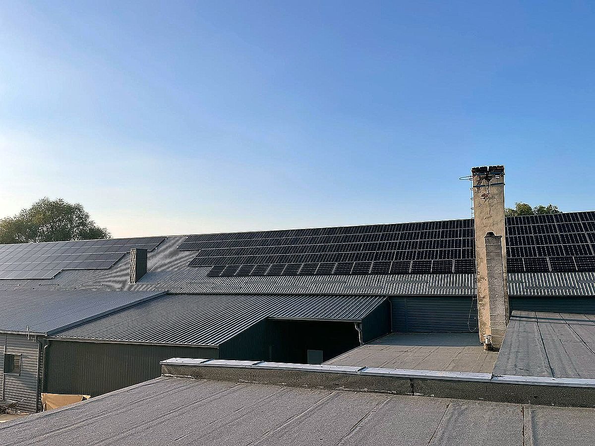 Instalacja fotowoltaiczna na dachu budynku przemysłowego z kominem. Panele ułożone rzędami, widok z góry na tle błękitnego nieba. Realizacja z wykorzystaniem OZE.