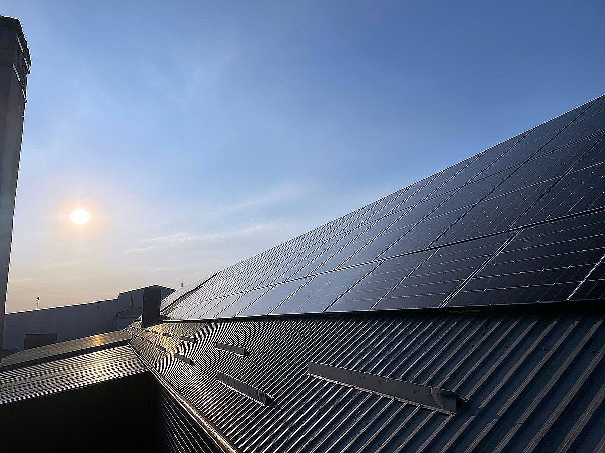 Panele słoneczne na metalowym dachu budynku w słoneczny dzień, widoczne kominy i błękitne niebo. Ujęcie z dołu.