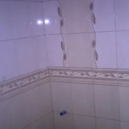 Wykończona ściana w łazience z jasnymi płytkami ceramicznymi, dekoracyjnym pasem z motywem liści i pionowym wzorem z brązowymi akcentami.