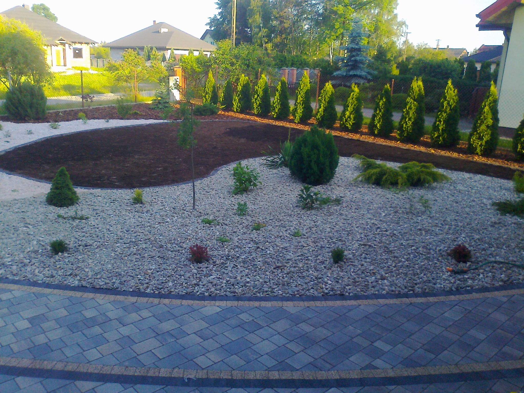 Nowo założony ogród z wysypanym żwirem i ziemią, obrzeżem z kostki brukowej oraz rzędem tuj przy ogrodzeniu.