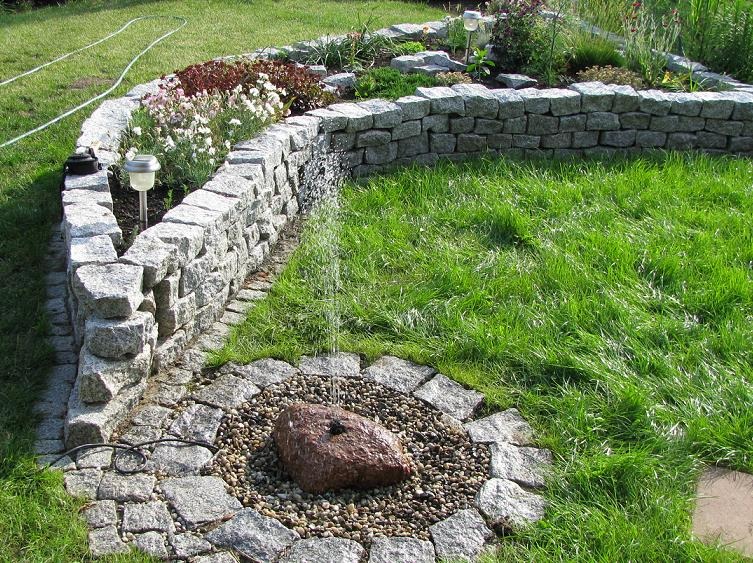 Ogród z kamienną rabatą w kształcie półkola i fontanną z kamienia otoczoną drobnymi kamykami, trawnik z zieloną trawą.
