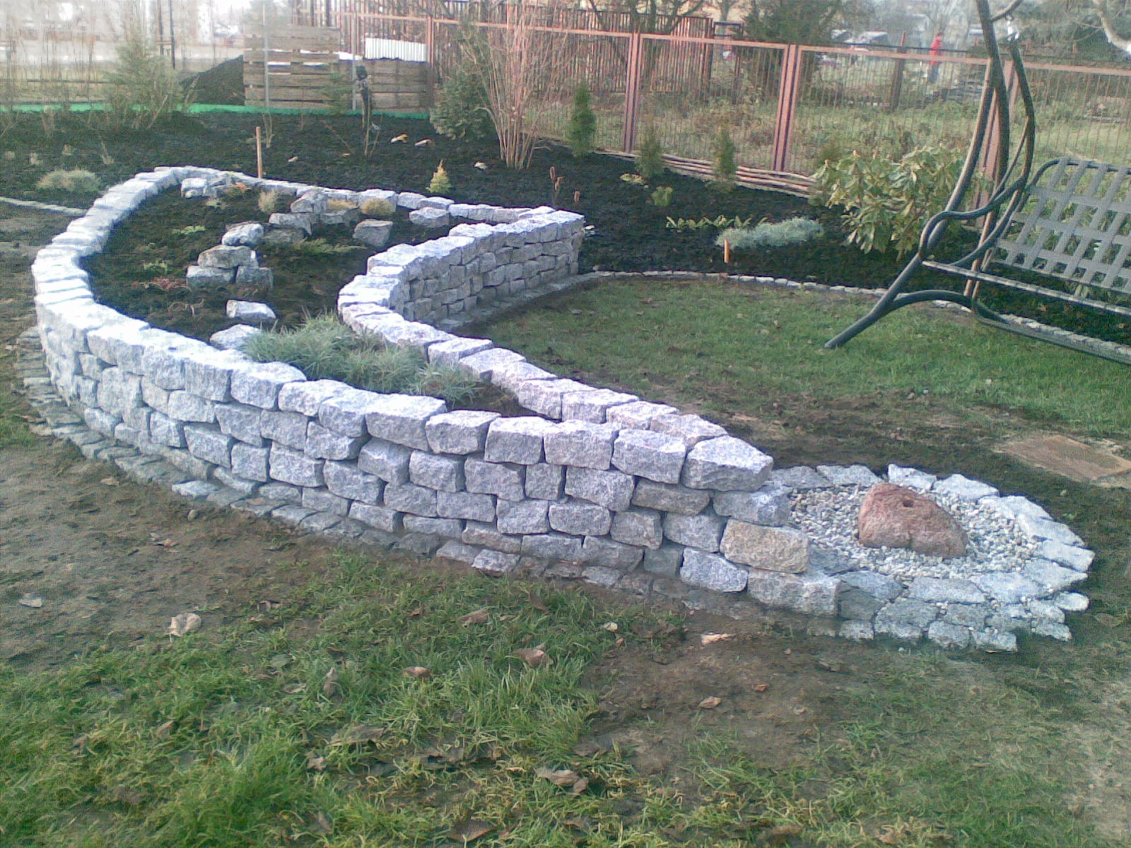 Kaskadowa rabata kwiatowa z kamienną obudową, wypełniona czarną korą i młodymi roślinami, widoczna huśtawka ogrodowa w tle.