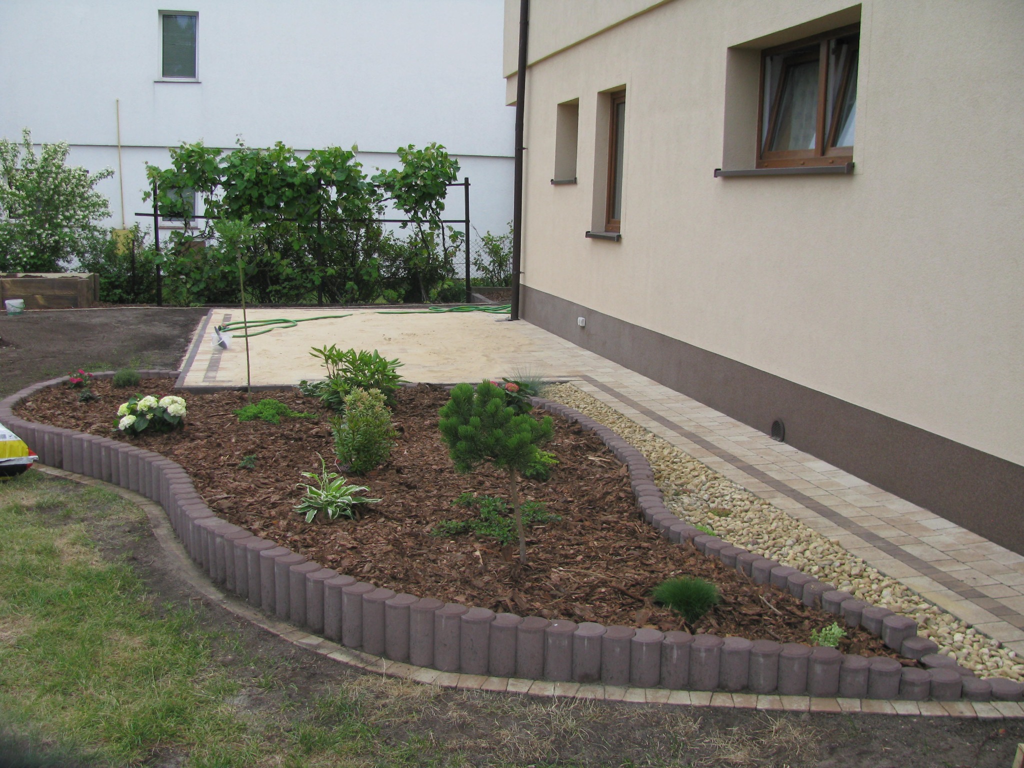 Nowo założony ogródek przed domem z obrzeżem z betonowych palisad, wysypany korą i obsadzony młodymi roślinami iglastymi oraz kwitnącymi bylinami, z widoczną ścieżką z kostki brukowej i fragmentem...