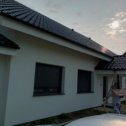 MIC-BUD - Biała elewacja domu jednorodzinnego z czarną dachówką, pracownik z deskami i wężem, słoneczny dzień. Widoczne okna z roletami zewnętrznymi i fragment dachu sąsiedniego budynku.