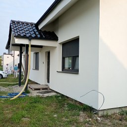 MIC-BUD - Biała elewacja domu jednorodzinnego z czarną dachówką, widoczni pracownicy w tle, żółty wąż do agregatu tynkarskiego. Świeżo położony tynk.