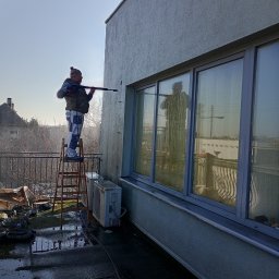 MIC-BUD - Pracownik na drabinie czyści elewację budynku myjką ciśnieniową. Widoczne okna z odbiciem sylwetki, mokry balkon i fragment sąsiedniego domu.