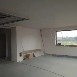 Wnętrze w stanie surowym: widoczne profile sufitowe, częściowo położona płyta karton-gips na suficie, gładkie ściany, narzędzia i materiały budowlane przy ścianie, okno z widokiem na zewnątrz.