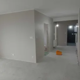 Wnętrze mieszkania w Czerniejewie po malowaniu ścian na szaro, widoczne gładkie powierzchnie, listwy przypodłogowe i ceramiczna podłoga. Pomarańczowe wiadro malarskie stoi w głębi pomieszczenia.