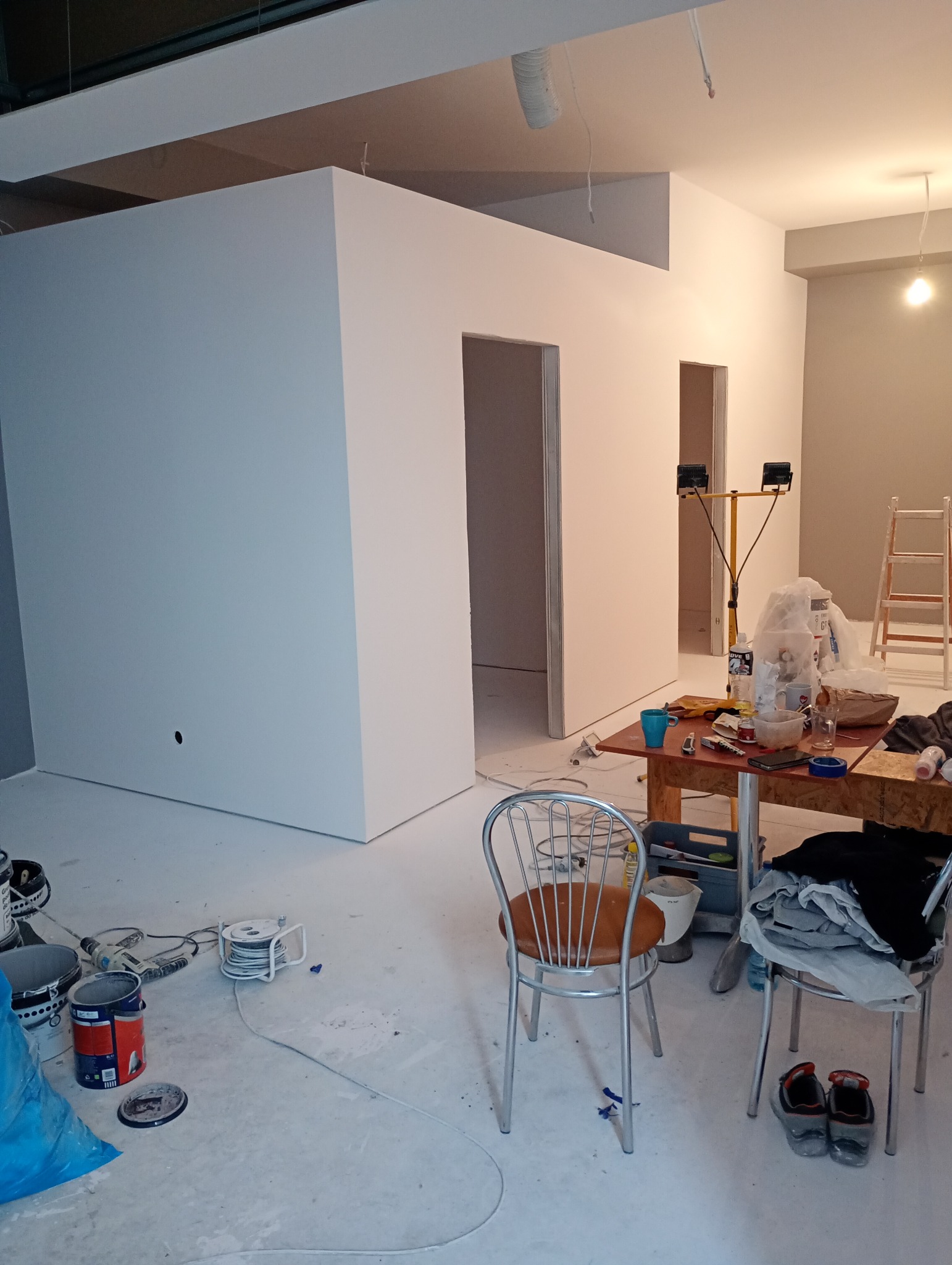 Wnętrze w trakcie prac wykończeniowych: świeżo pomalowane ściany na biało, widoczne narzędzia malarskie, kable na podłodze, stół z akcesoriami i lampa budowlana. Dwa krzesła stoją na pierwszym planie.