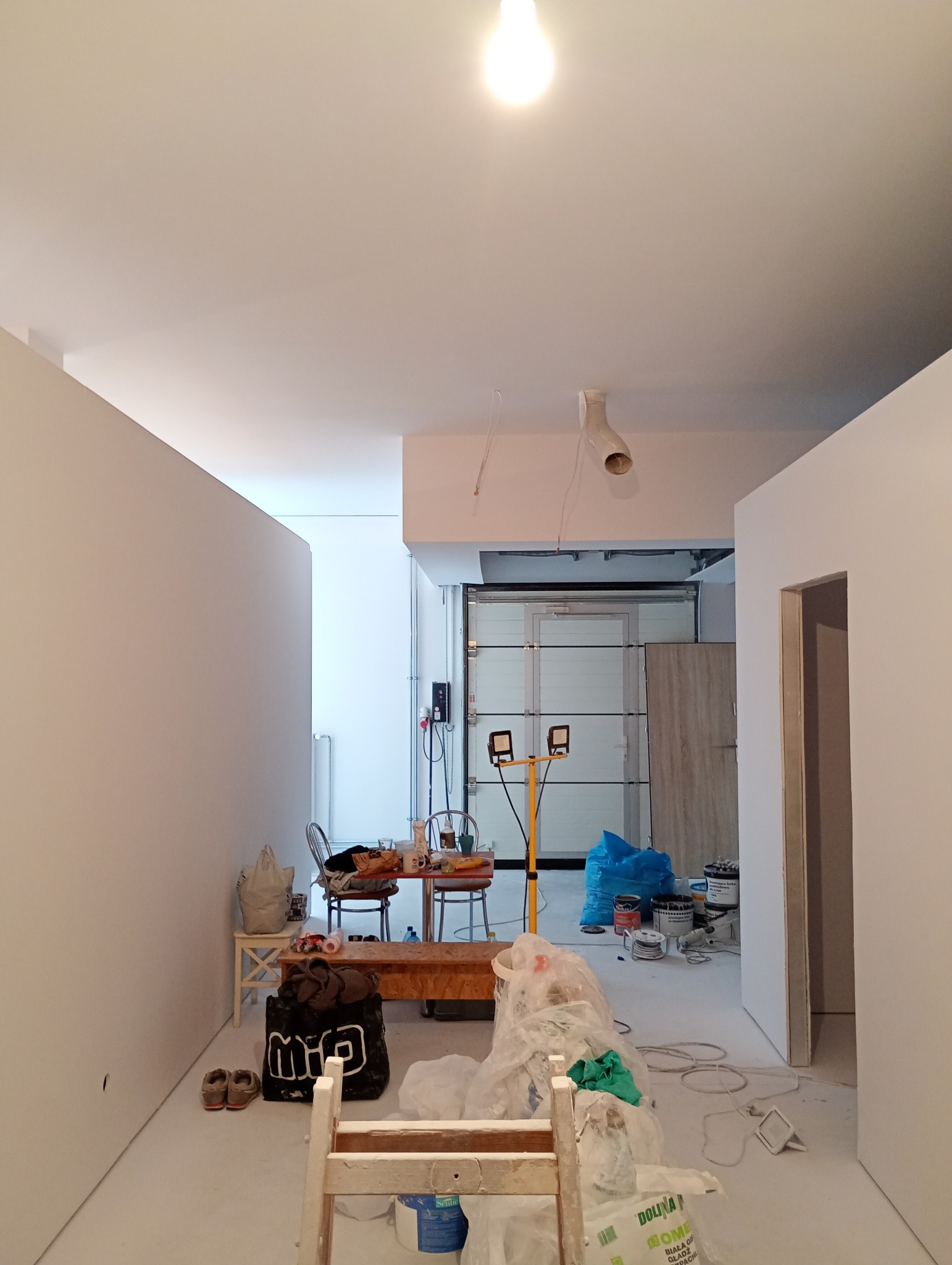 Wnętrze w trakcie remontu: białe ściany, prowizoryczny stół z krzesłami, lampy budowlane, worki z materiałami budowlanymi i narzędzia malarskie.