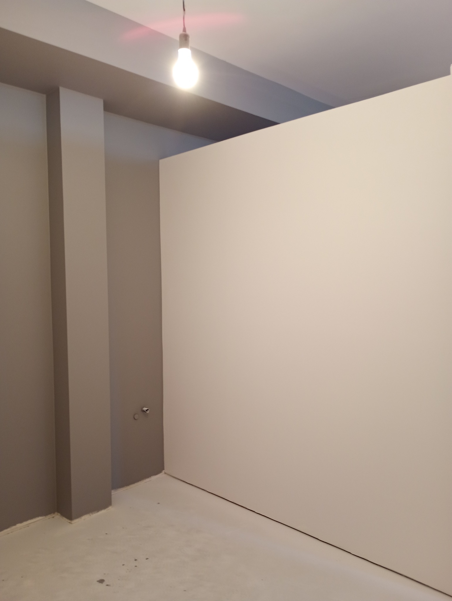 Wnętrze w trakcie prac wykończeniowych: ściana pomalowana na biało, szara kolumna i sufit, widoczna żarówka sufitowa.