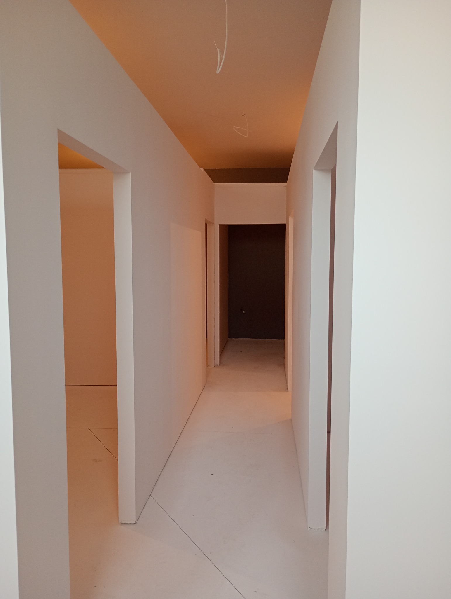 Korytarz w stanie surowym, ściany i sufit pomalowane na biało, widoczne przewody elektryczne wystające z sufitu, przygotowane do dalszych prac wykończeniowych.