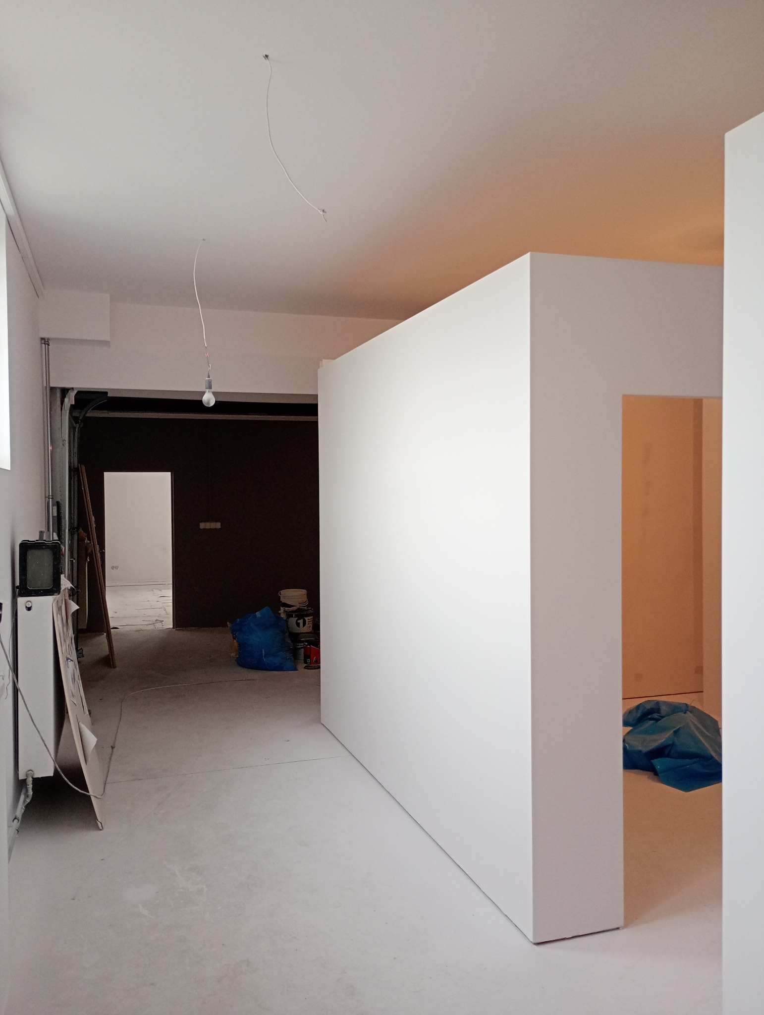 Wnętrze w trakcie remontu: białe ściany działowe, surowy beton na podłodze, wisząca żarówka bez klosza, worki z materiałami budowlanymi w tle.