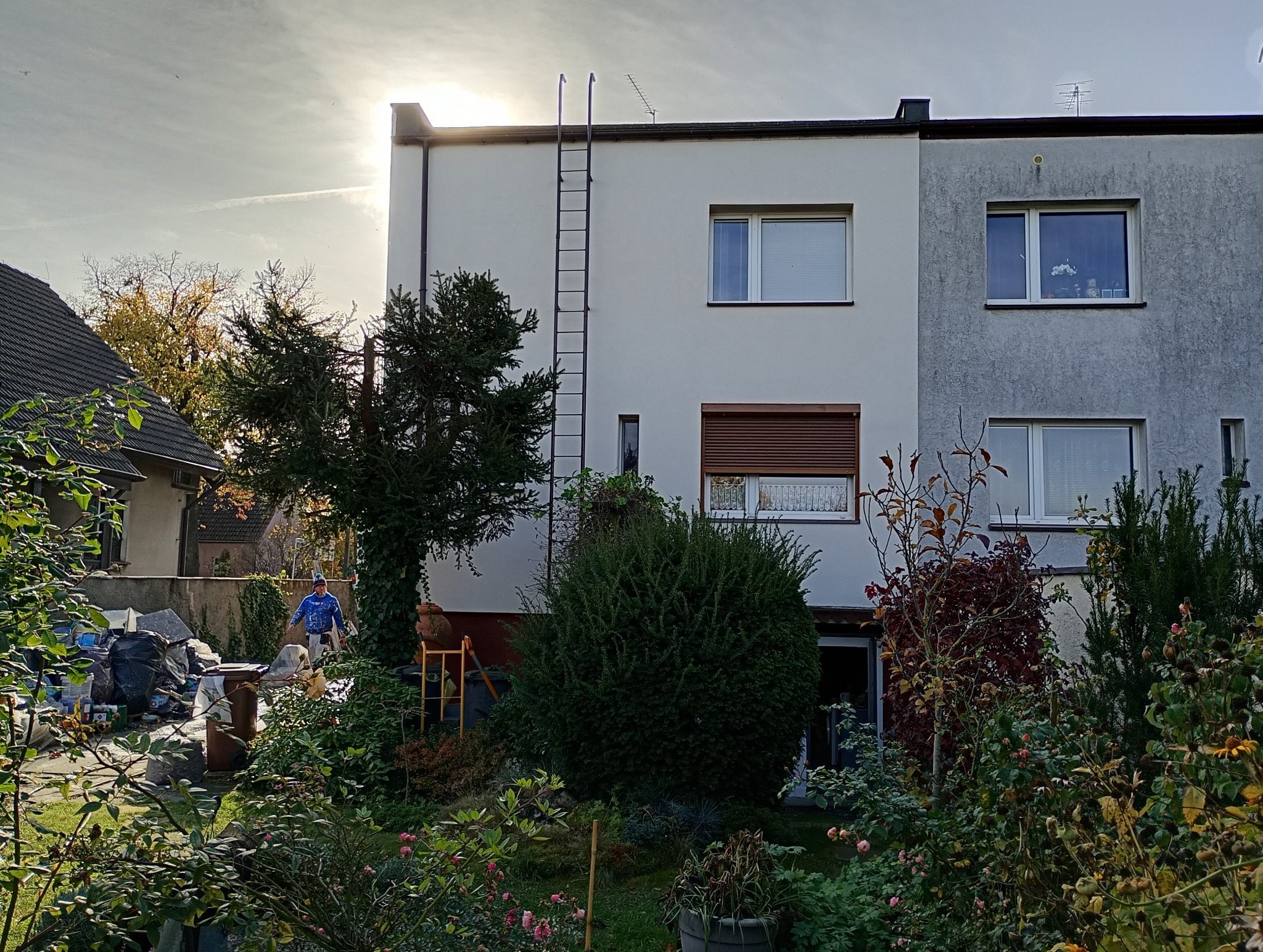 Dwukondygnacyjny dom z drabinką na elewacji, widoczny zza bujnej zieleni ogrodu. Osoba w roboczym ubraniu w tle, sugerująca prace remontowe.