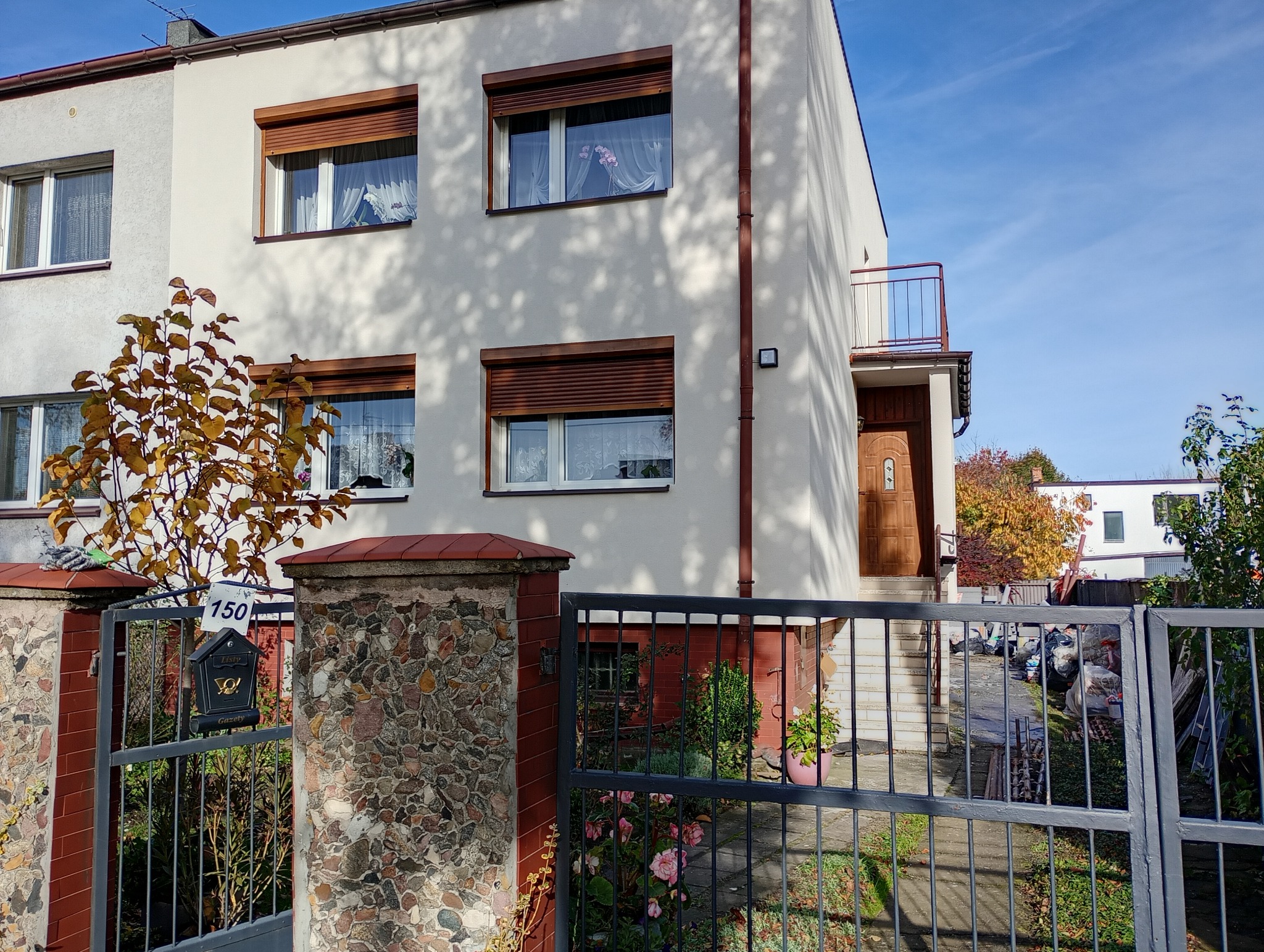 Dwukondygnacyjny dom z jasną elewacją i brązowymi roletami okiennymi, widziany zza metalowego ogrodzenia i kamienno-ceglanego słupka z numerem 150.