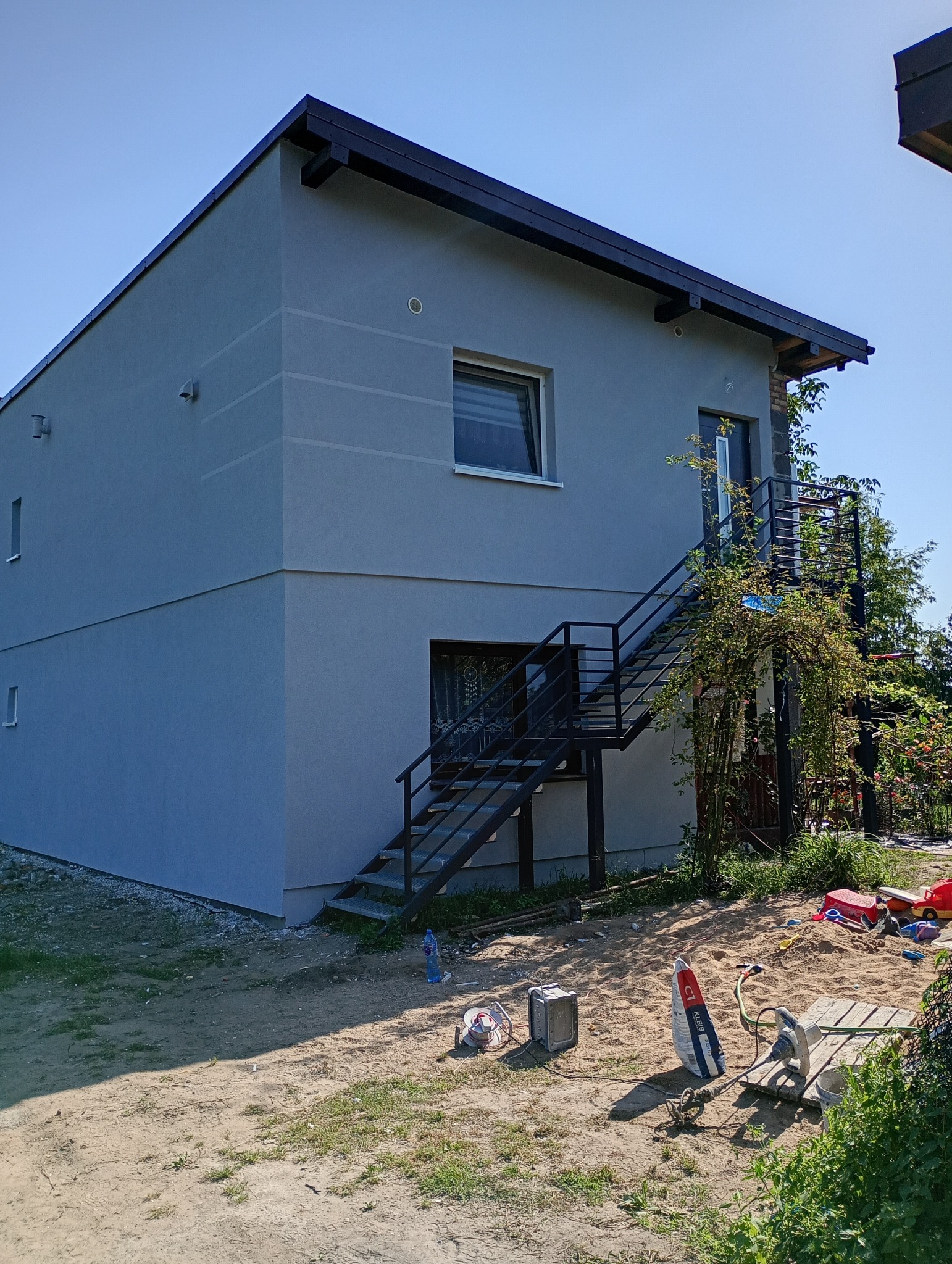 Dwukondygnacyjny dom z szarą elewacją i zewnętrznymi schodami. Widoczne narzędzia i materiały budowlane na trawniku. Błękitne niebo w tle.